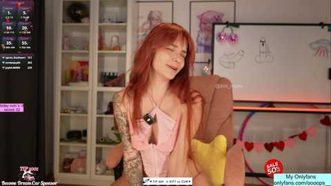 Video of a pale-skinned, red-haired woman in a pink halter top, sitting in a cozy room with shelves, heart decorations, and a laptop screen visible.