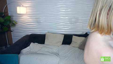 Video of a blonde woman from behind, standing beside a wavy, white-textured wall in a modern living room with a dark sofa, beige pillows, and a green plant in the corner.