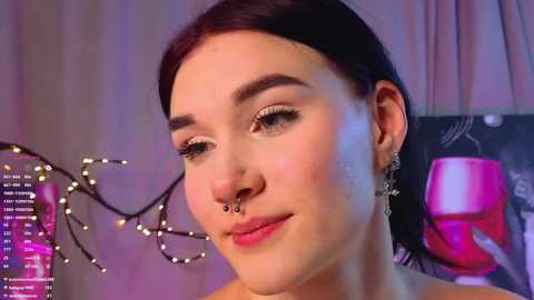 Video of a young woman with fair skin, dark hair, and a septum piercing, wearing a pink top. She has a soft smile and a subtle makeup look. The background features a pink cup and string lights.