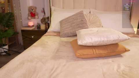 Video of a neatly made bed with beige and brown pillows, plush teddy bear, and wooden nightstand in a cozy, warmly lit bedroom.