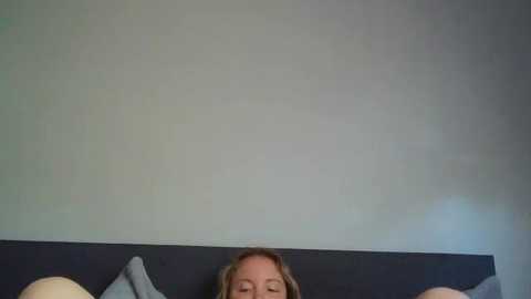 Video of a woman lying on a dark bed, her face partially visible, with a light-colored pillow and a gray wall in the background.