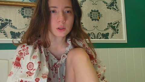Video of a young girl with long brown hair, fair skin, and wearing a floral-patterned dress, sitting against a green and cream wallpaper with intricate designs.