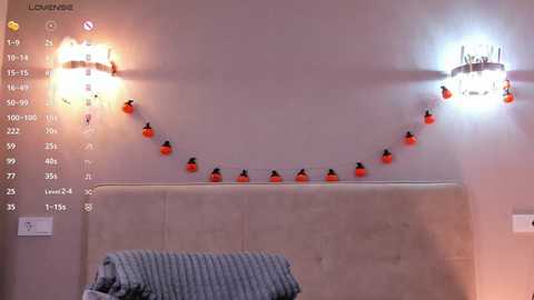 Video of a cozy, dimly lit bedroom with beige walls, a tufted headboard, and a cozy throw blanket. Decorated with warm, glowing string lights and two wall sconces.