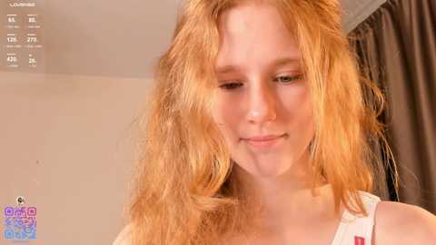 Media: Video of a young Caucasian woman with long, wavy, light brown hair, wearing a white tank top, smiling gently. Background features beige walls and brown curtains.