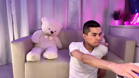 A video of a young man with short dark hair in a white t-shirt, sitting on a beige couch with two plush white teddy bears, under pink and purple lights.