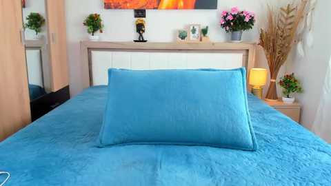 Media: Video of a neatly made bed with a blue quilt and matching pillow, set against a light wooden headboard, white dresser, and colorful floral artwork on a white wall.