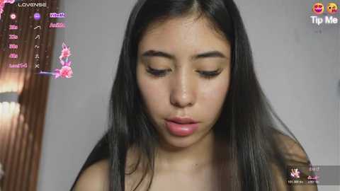 A video of a young woman with long, straight black hair and light skin, wearing a black top, looking down with a contemplative expression. Background includes a blurred, plain wall and a partially visible wooden object.