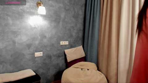 Video of a minimalist bedroom with gray textured walls, beige bedspread, and a white wall-mounted light. A person in a red dress stands in the background.