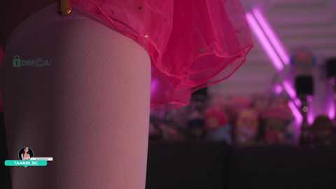 Media: A close-up video of a light-skinned person wearing a bright pink tutu, with blurred background of a crowded room.