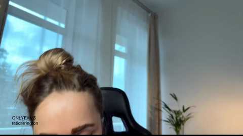 Video of a blonde woman with a messy bun, partially visible, in a modern office with a black chair, plants, and large windows with sheer curtains.