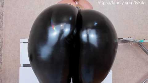 Video of a person in a shiny, black latex bodysuit, highlighting their large, rounded buttocks and thighs. The background shows a tiled wall and a medical device.