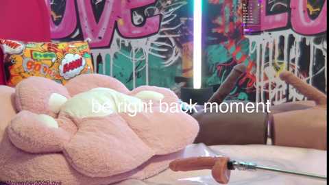 Media: Video of a plush pink flamingo on a bed, surrounded by vibrant graffiti art and a \"LOVE\" sign, with the text \"be right back moment\" overlayed.