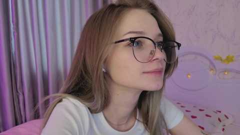 Video of a fair-skinned, young woman with long, blonde hair, wearing glasses, and a white t-shirt, sitting against a light purple, floral wallpaper background.