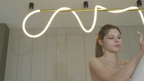 Video of a nude woman with fair skin and brown hair, applying makeup in a modern bathroom with white cabinets and a neon sign above her.