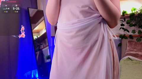 A video of a woman in a white, sheer dress, standing in a room with a large flat-screen TV showing a cartoon, a green plant, and a couch.