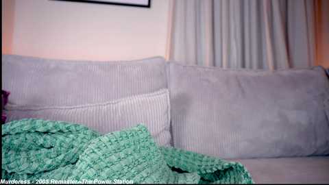 Video of a beige couch with light green knitted blanket and white curtains, soft lighting, simple decor, and a framed picture partially visible.