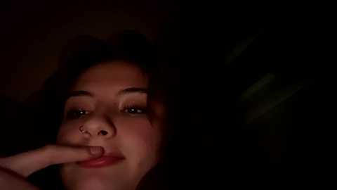 A dimly lit video of a young woman with dark curly hair, medium skin tone, and light makeup, biting her finger in a close-up, intimate setting.