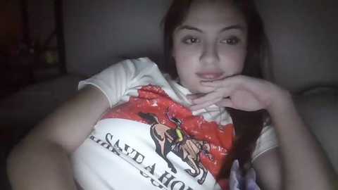 Video of a young woman with light skin, dark hair, and brown eyes, wearing a white t-shirt with a graphic design, seated on a couch, hand on her chin.