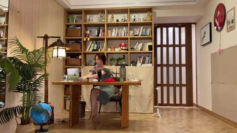 Video of a cozy, cluttered study with wooden bookshelves, a potted plant, a desk, and a woman artist working on a painting.