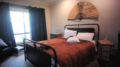 Video of a minimalist bedroom with a single bed, orange bedding, black metal frame, white pillows, and a decorative fan on the wall.