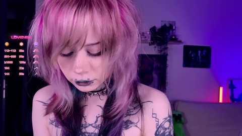 Video of a pale-skinned young woman with pink and purple hair, wearing dark lipstick and a black choker, with intricate black tattoos covering her chest and arms. She stands in a dimly lit room with purple and blue lighting, a bookshelf, and a digital clock in the background.