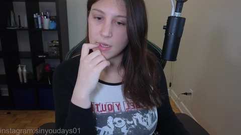 A video of a young woman with fair skin, long brown hair, and wearing a black hoodie with a graphic design. She sits in an office chair, biting her fingernail, in a minimalist room with wooden floors and a bookshelf.