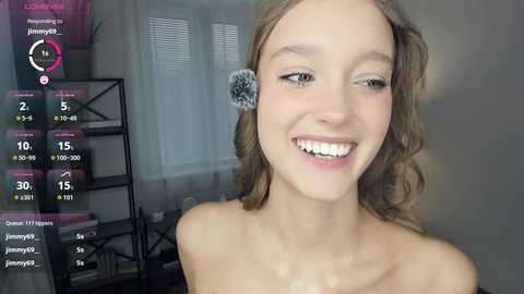 Media: Video of a smiling, light-skinned young woman with wavy blonde hair, wearing a white top, in a modern room with a black shelf and blinds.