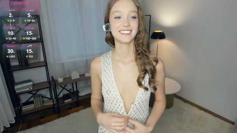 Video of a young, fair-skinned girl with long, wavy blonde hair, wearing a white crochet halter dress and a headset, smiling in a modern, well-lit living room.