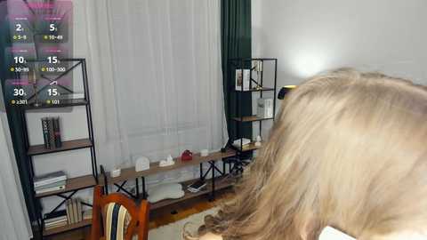 Media: Video of a woman with long blonde hair, playing a video game, in a modern living room with a TV, bookshelf, and white curtains.