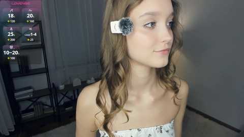 Media: A video of a young woman with fair skin, wavy brown hair, and a white floral strapless top. She's in a dimly lit room, with a shelf filled with books and a window in the background.