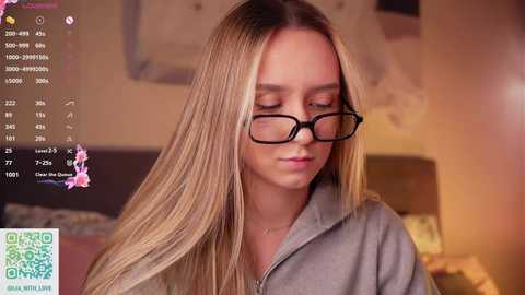 A video of a young Caucasian woman with long, straight blonde hair, wearing black-rimmed glasses and a grey hoodie, gazing downward in a cozy indoor setting.