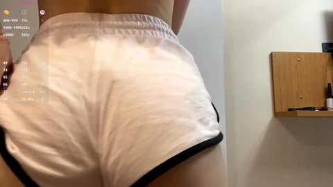 Media: Video of a woman's lower back and buttocks in tight, white athletic shorts with black trim, taken indoors with a wooden shelf and beige wall in the background.