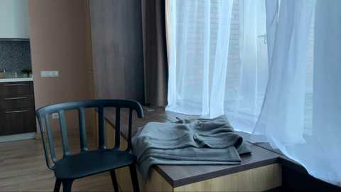 A video of a minimalist room with a dark wooden chair and a gray blanket draped over a wooden bench near a window with sheer white curtains.