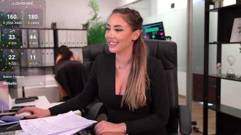 Video of a smiling, light-skinned woman with long, brown hair in a ponytail, wearing a black, low-cut dress, sitting at a desk in a modern office with computer screens and bookshelves in the background.