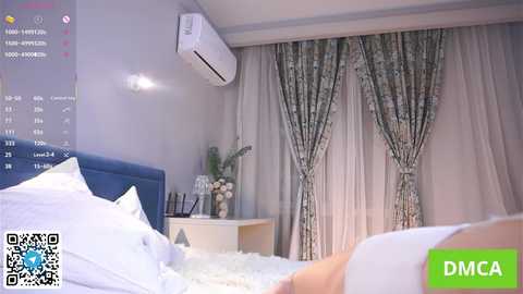 Video of a modern bedroom with a blue upholstered headboard, white bedding, an air conditioner on the wall, and a window with floral curtains.
