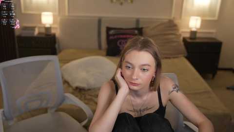 A video of a young woman with fair skin and shoulder-length blonde hair, wearing a black spaghetti strap top, sitting on a bed in a cozy bedroom with beige walls, white lamps, and a floral pillow.