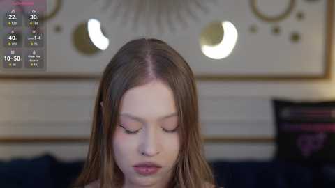 Video of a young woman with long, straight brown hair, wearing makeup, with closed eyes, standing in front of a boho-style wall art, in a dimly lit room.