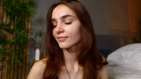 Video of a young woman with long brown hair, closed eyes, and a serene expression, wearing a black necklace. Background features a blurred bamboo screen and a bed with white sheets.