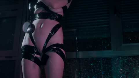 Video of a person in black, shiny latex bondage gear, standing in a dimly lit room with a glittery wall.