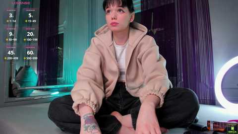 Video of a young, fair-skinned woman with short black hair, wearing a beige hoodie and black pants, sitting cross-legged in a modern, dimly lit room with purple curtains and a circular neon light.