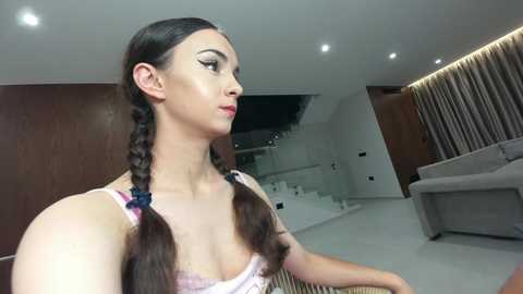 Video of a light-skinned woman with long brown hair in braids, wearing a white lace bra, standing in a modern living room with minimalist decor.