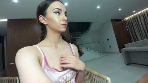 Video of a fair-skinned woman with dark braided hair, wearing a pink camisole, standing in a modern, minimalist room with white walls, wooden furniture, and a staircase.