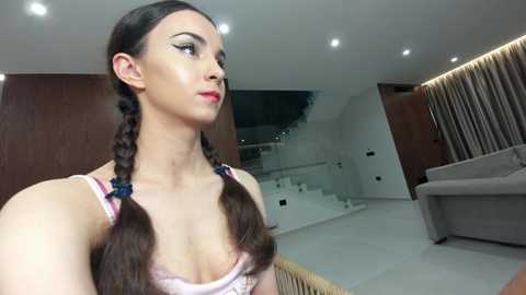 Video of a slim, fair-skinned woman with long brown hair styled in twin braids, wearing a light pink lace bra. She gazes thoughtfully to the left in a modern, minimalist living room with white walls, wooden accents, and a beige couch.