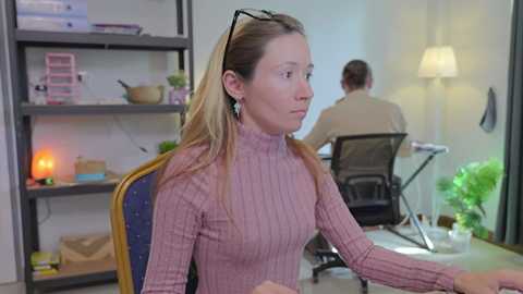 Media: Video of a blonde woman in a pink sweater, seated at a desk in a modern office, with shelves and plants in the background.