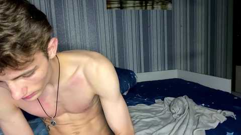 Video of a shirtless, muscular, fair-skinned young man with short brown hair, wearing a black necklace, lying on a bed with blue sheets in a dimly lit room with vertical striped wallpaper.