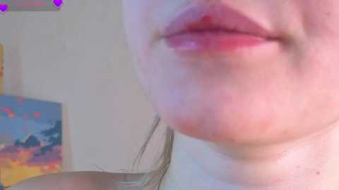 Close-up video of a Caucasian woman's face with red lips, slightly smiling, showing puffy cheeks and a hint of a pinkish nose. Background features a blurred wall with a colorful abstract painting.