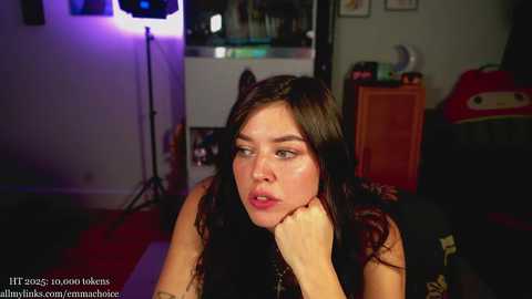 Video of a young woman with long, wavy dark hair, resting her chin on her hand, in a dimly lit room with a red carpet, a studio light, and various items in the background.