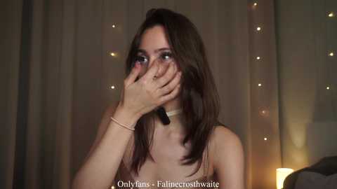 A video of a young woman with fair skin and long dark hair, wearing a beige top, covering her face with her hands in a dimly lit room with string lights.