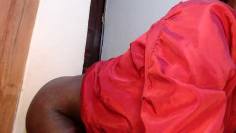 Video of a person with medium brown skin, wearing a bright red dress, leaning against a wooden door frame. The background features a white wall.