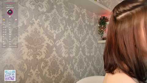 Media: Video of a woman with shoulder-length, straight, dark brown hair, sitting in a room with light gray, damask-patterned wallpaper and a small potted plant on a white shelf.